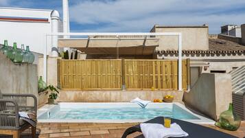 Un área de piscina en la azotea con cerramiento de madera, una silla de descanso, una mesita con un vaso de zumo de naranja y una botella de agua.