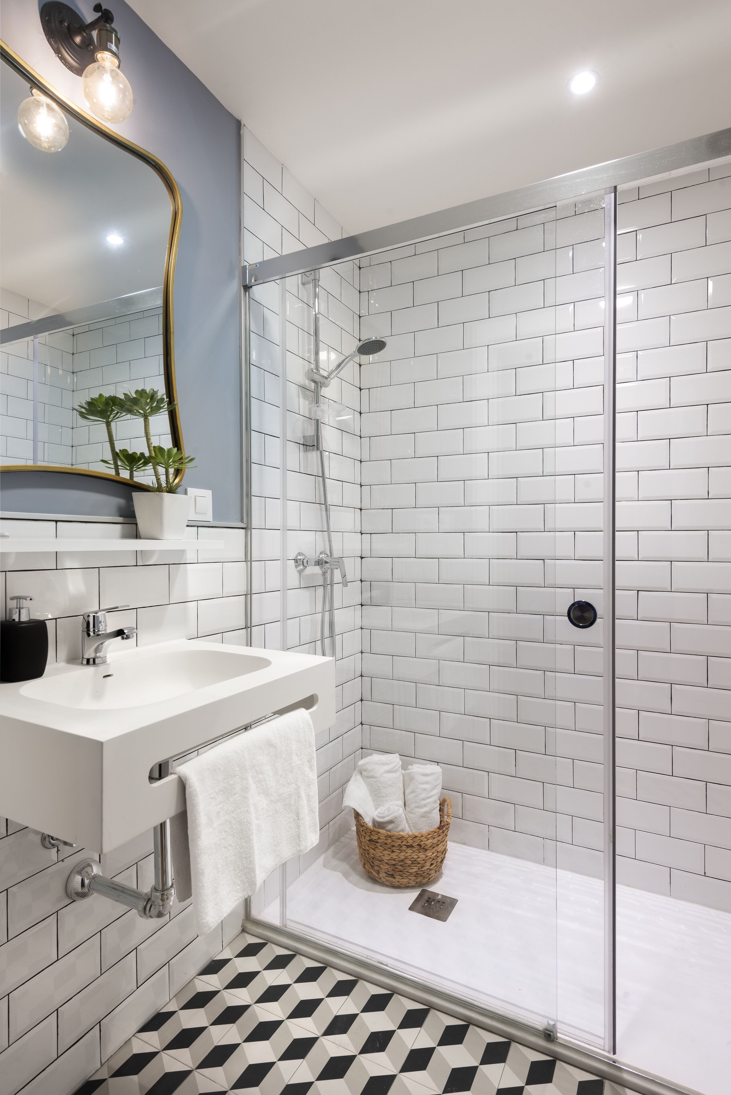 Une salle de bain moderne avec une douche à l’italienne en verre, des murs recouverts de carreaux blancs, un lavabo blanc avec un miroir et un sol à motifs.