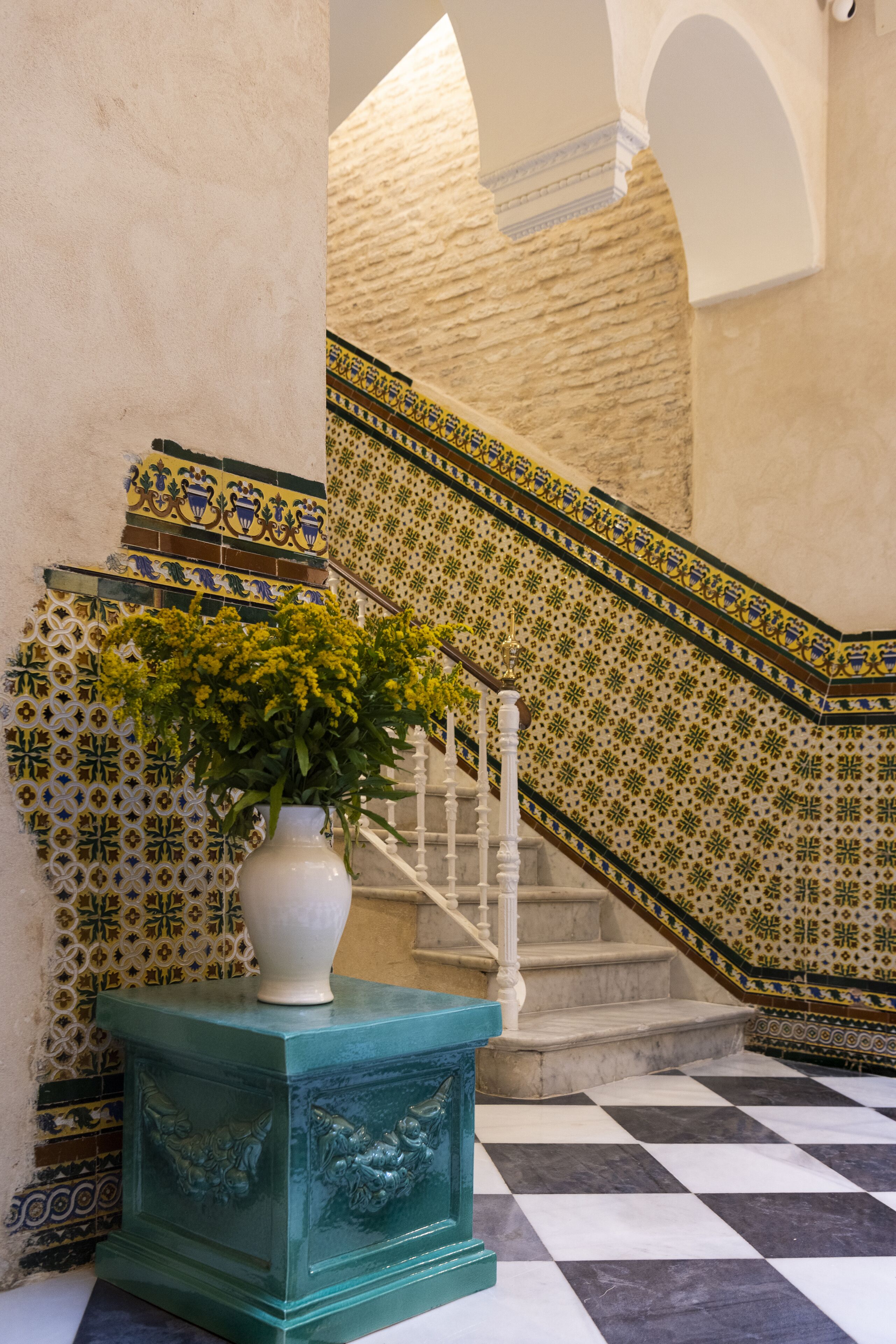 A staircase with patterned tiles, a white vase with yellow flowers, and a checkered floor.