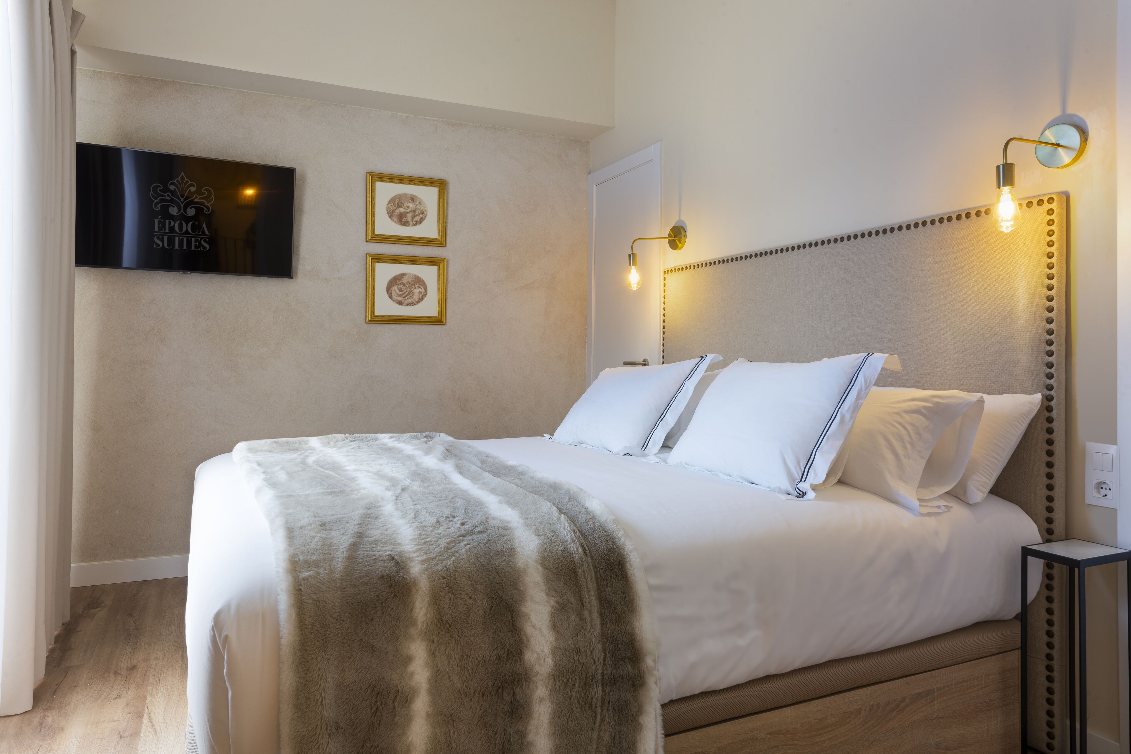 A neatly made bed with a fluffy blanket, a television mounted on the wall, and two framed pictures.