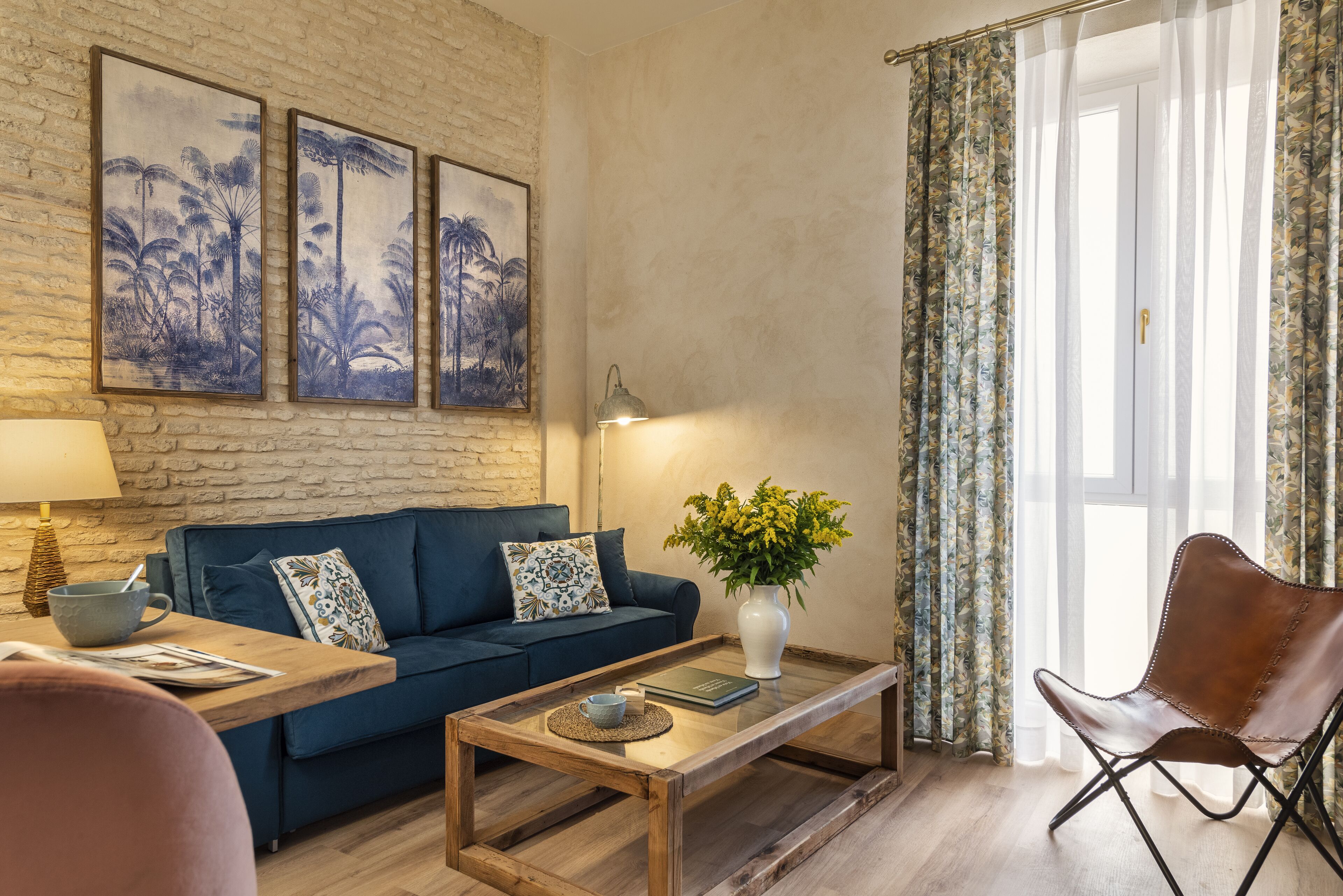 A living room with a blue sofa, a wooden coffee table, a vase with flowers, and a chair. There are framed pictures on the wall and a lamp on the side table.