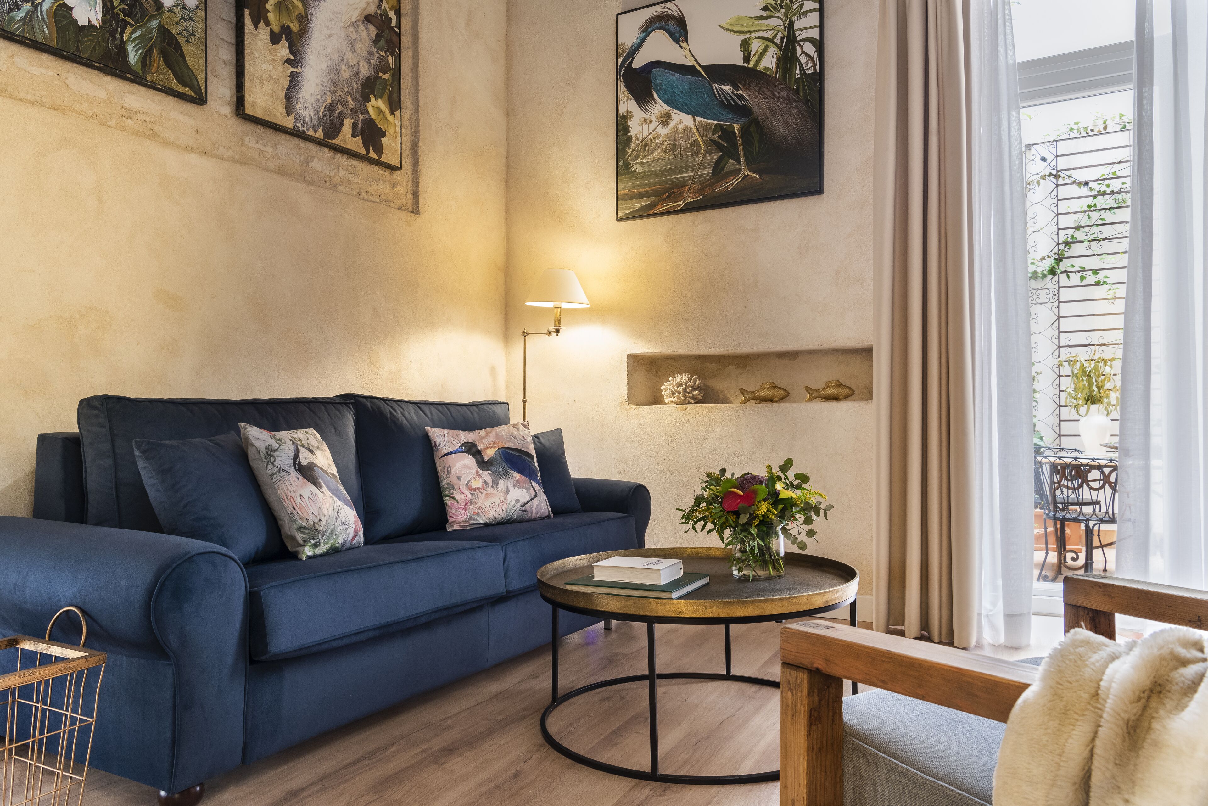 Een woonkamer met een donkerblauwe bank, een rond bijzettafeltje met een boek en bloemen, een lamp en wanddecoratie.