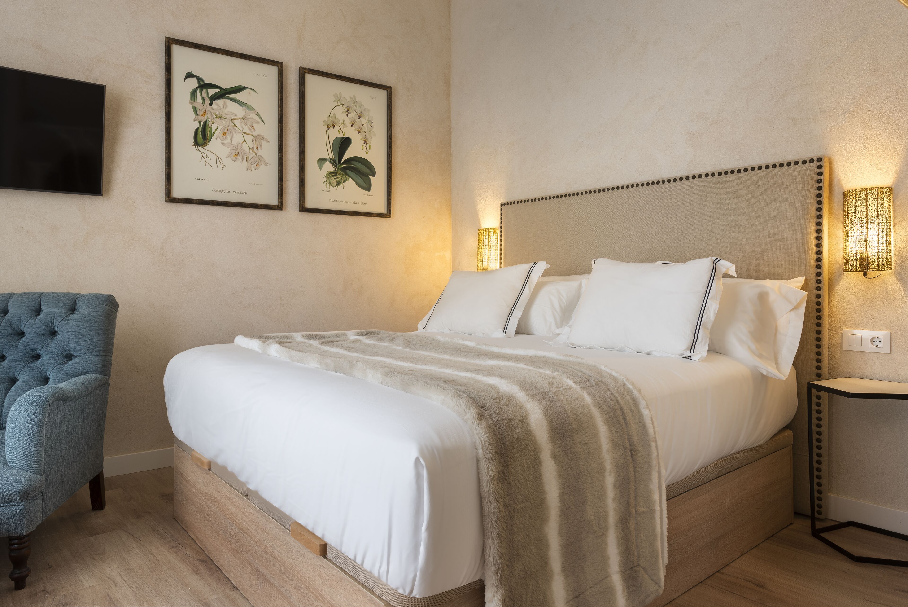 Un lit bien fait, avec du linge de lit blanc et un plaid beige, un fauteuil bleu recouvert de tissu et deux reproductions botaniques encadrées accrochées au mur.
