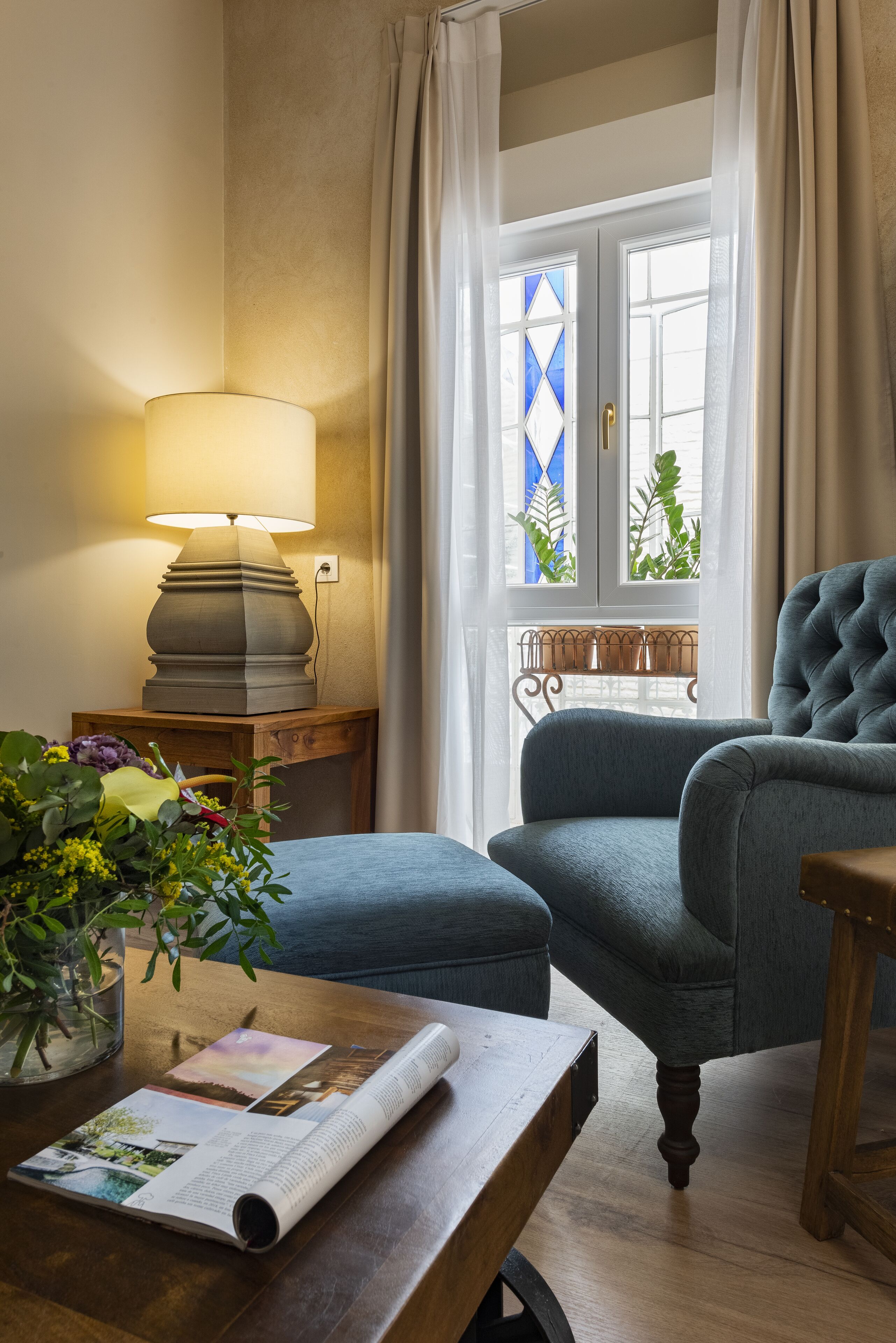 A room with a wooden side table, a lamp, a blue armchair, a window with curtains, and a view of greenery outside.