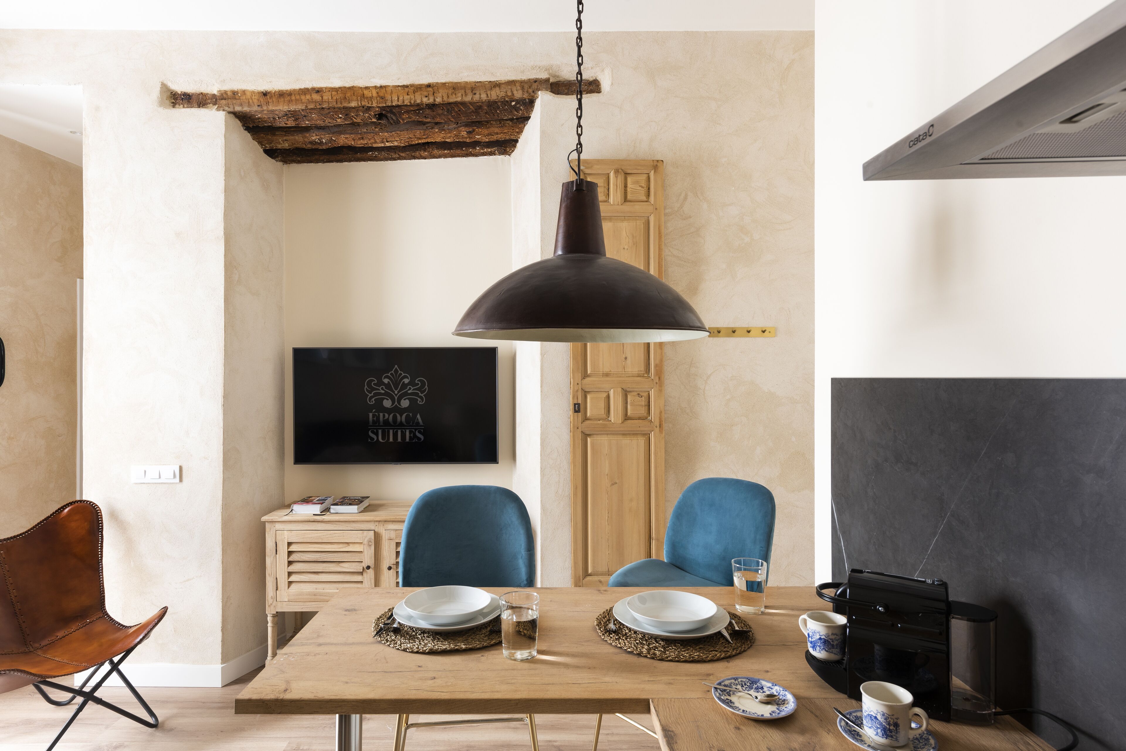 A dining area with a wooden table, blue chairs, a hanging pendant light, and a television on the wall.