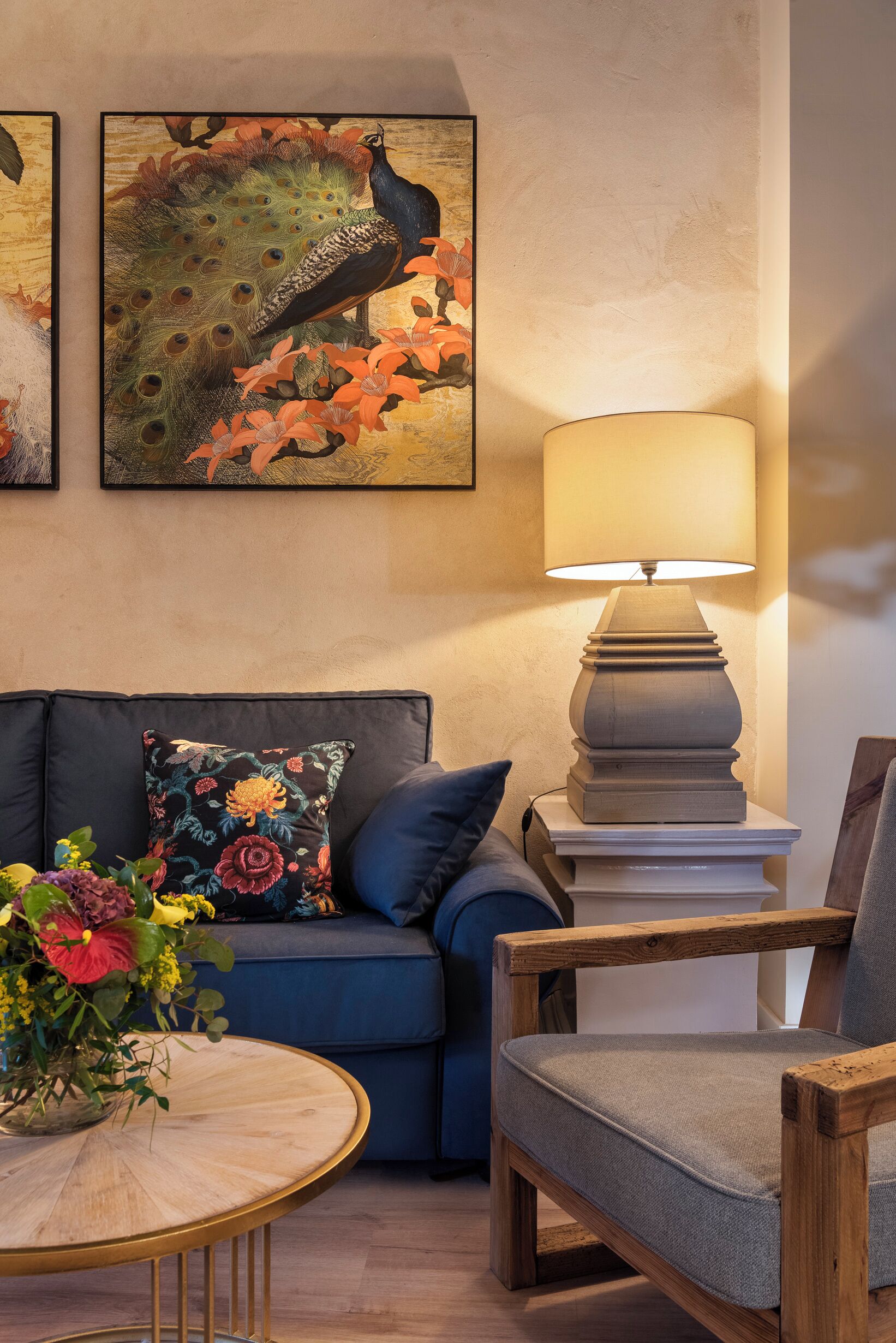 A living room with a blue sofa, a wooden chair, a round wooden table with a vase of flowers, and two framed artworks on the wall.