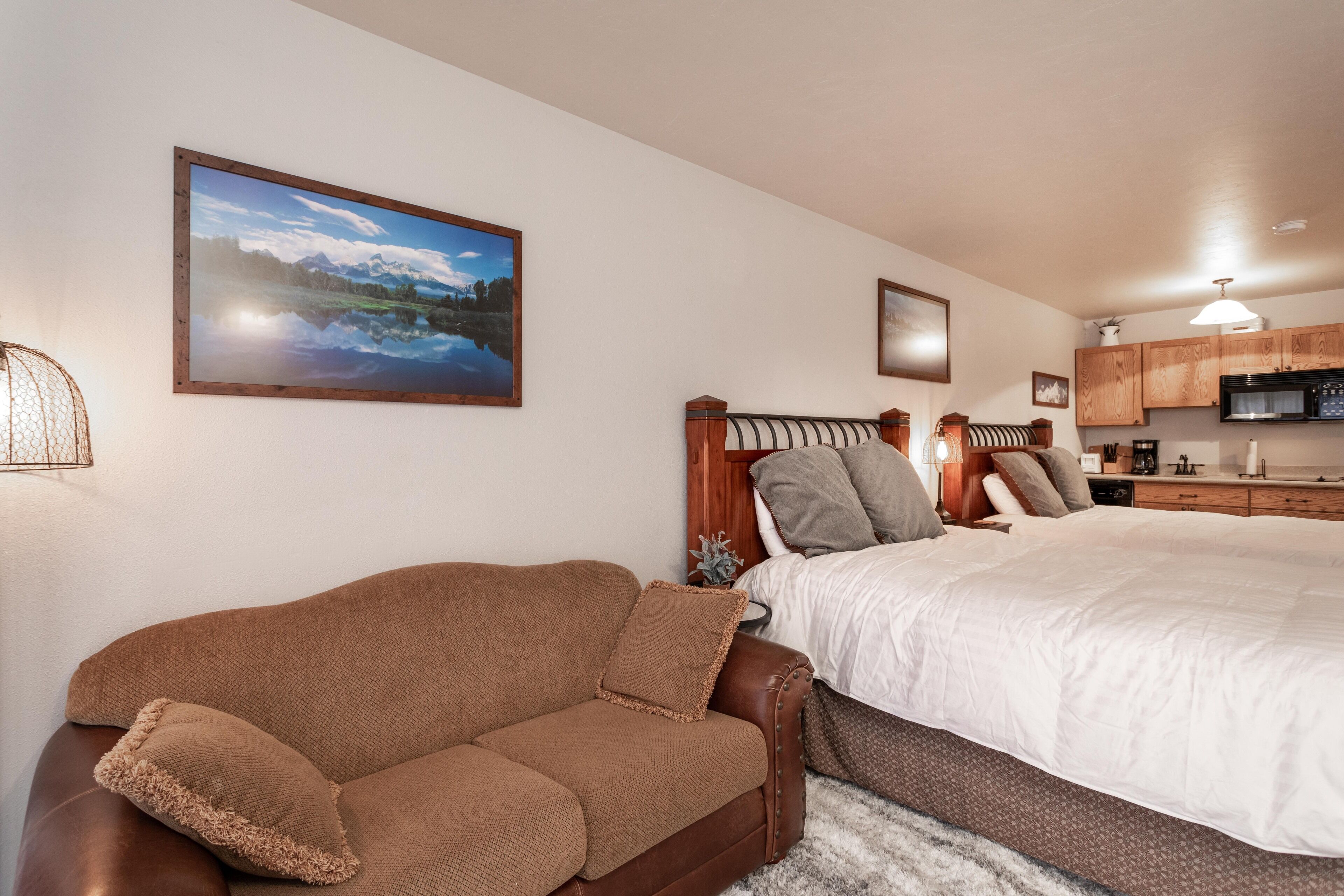 A bedroom with a bed, a brown leather sofa, a wooden headboard, and a kitchen area with wooden cabinets and a microwave.
