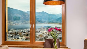 Uma sala de jantar com uma mesa, cadeiras e um vaso com flores. Uma janela grande oferece vista para um lago e montanhas.
