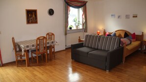 A living room with a wooden floor, a sofa, a dining table with chairs, a window with curtains, and a radiator. - Pension & Ferienwohnung Breitmoser (Riedenburg)