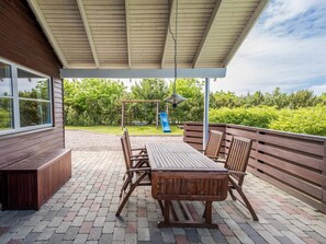 Un patio exterior con mesa y sillas de madera, un columpio y un tobogán, rodeado de follaje.