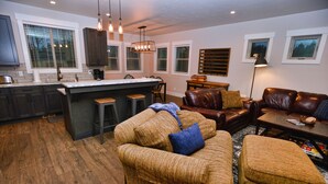 Een ruime woonkamer met een bruine bank, voetenbankje en een salontafel. Er is een keuken met een groot eiland en barstoelen. De kamer heeft een houten vloer en grote ramen met zonwering.
