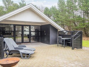Ein überdachter Außenbereich mit Tisch und Stühlen, einem Grill und einem Gebäude mit großen Glasfenstern.