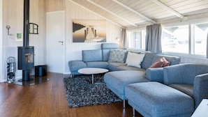 A modern living room with a built-in wood stove, a grey sectional sofa, a round coffee table, and a large window with curtains. - Bright Dune View Cottage-by Traum (Hvide Sande)