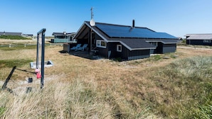 A modern, single-story building with a sloped roof, a satellite dish, and a small porch with two chairs. - Bright Dune View Cottage-by Traum (Hvide Sande)