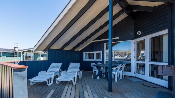 A covered outdoor deck with lounge chairs and a table, surrounded by a wooden deck and a dark blue wall. - Bright Dune View Cottage-by Traum (Hvide Sande)