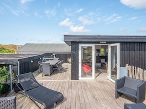 A wooden deck with outdoor furniture, a glass door leading to a kitchen, and a view of the surrounding landscape.