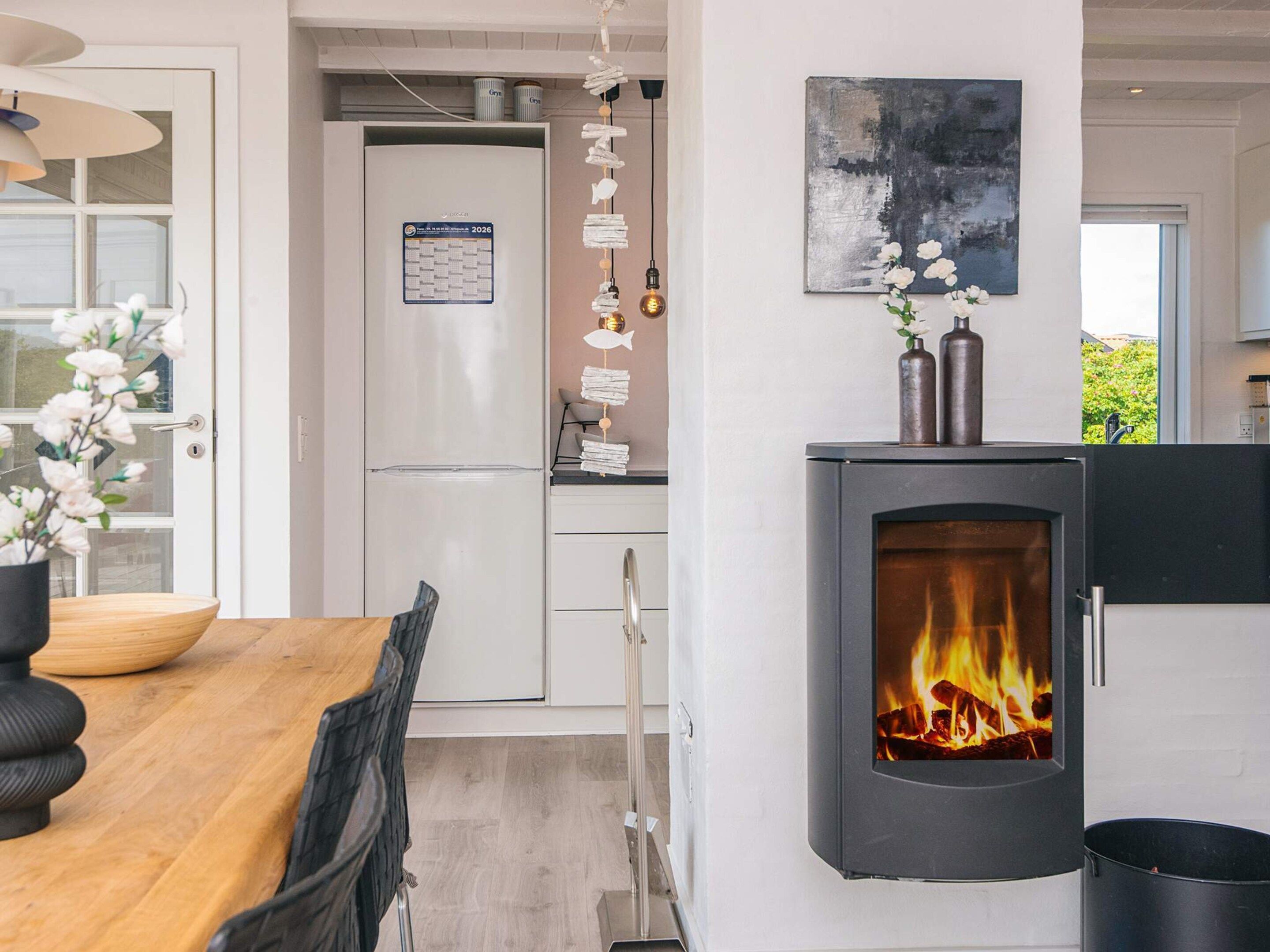 A modern kitchen with a built-in wood stove, a dining area with a wooden table and chairs, and a refrigerator with a calendar.