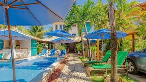 A pool area with lounge chairs and umbrellas, a stone pathway, and a building in the background. - GB19 Casa 7 Quartos a 50m da Praia (Camaçari)