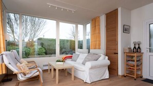 A modern living room with a white sofa, a wooden rocking chair, a small table, and a wooden shelf with bottles. - Quiet Home with Garden near Beach & Center (Renesse)
