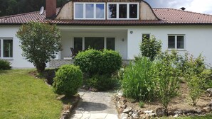 Une maison à deux étages, avec un toit en tuiles, un chemin de pierre menant à l’entrée et un jardin bien entretenu agrémenté de divers arbustes et plantes.