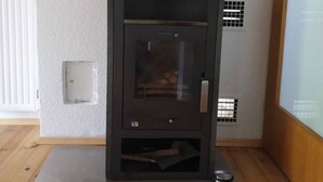 Un poêle à bois moderne, doté d’une porte en verre, est placé sur un sol gris foncé, adossé à un mur blanc sur lequel se trouve une petite étagère.
