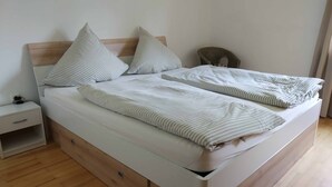 Un lit double avec une literie à rayures et deux oreillers, une table de chevet en bois dotée d'une prise électrique, et une petite chaise en osier.