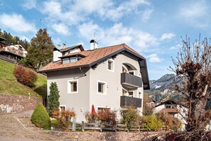 Een huis met twee verdiepingen, een schuin dak en een stenen trap, met op de achtergrond een uitzicht op bergen.