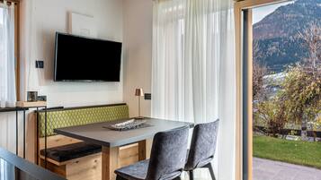 Ein modernes Zimmer mit Esstisch, Stühlen, einem flachbildfernseher an der Wand und einem großen Fenster mit Blick auf die Berge.