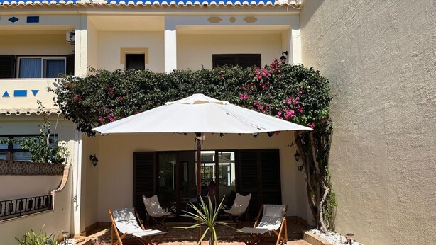 Een terras met twee loungestoelen en een grote parasol, omgeven door een betegelde vloer en potplanten.