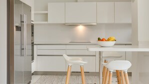 Una cocina moderna con armarios blancos, un refrigerador de acero inoxidable, un cuenco de fruta en la encimera y dos taburetes de bar blancos.