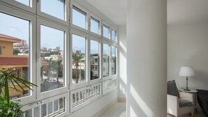 Una habitación con ventanas amplias, una silla, una lámpara y vistas a edificios y palmeras.