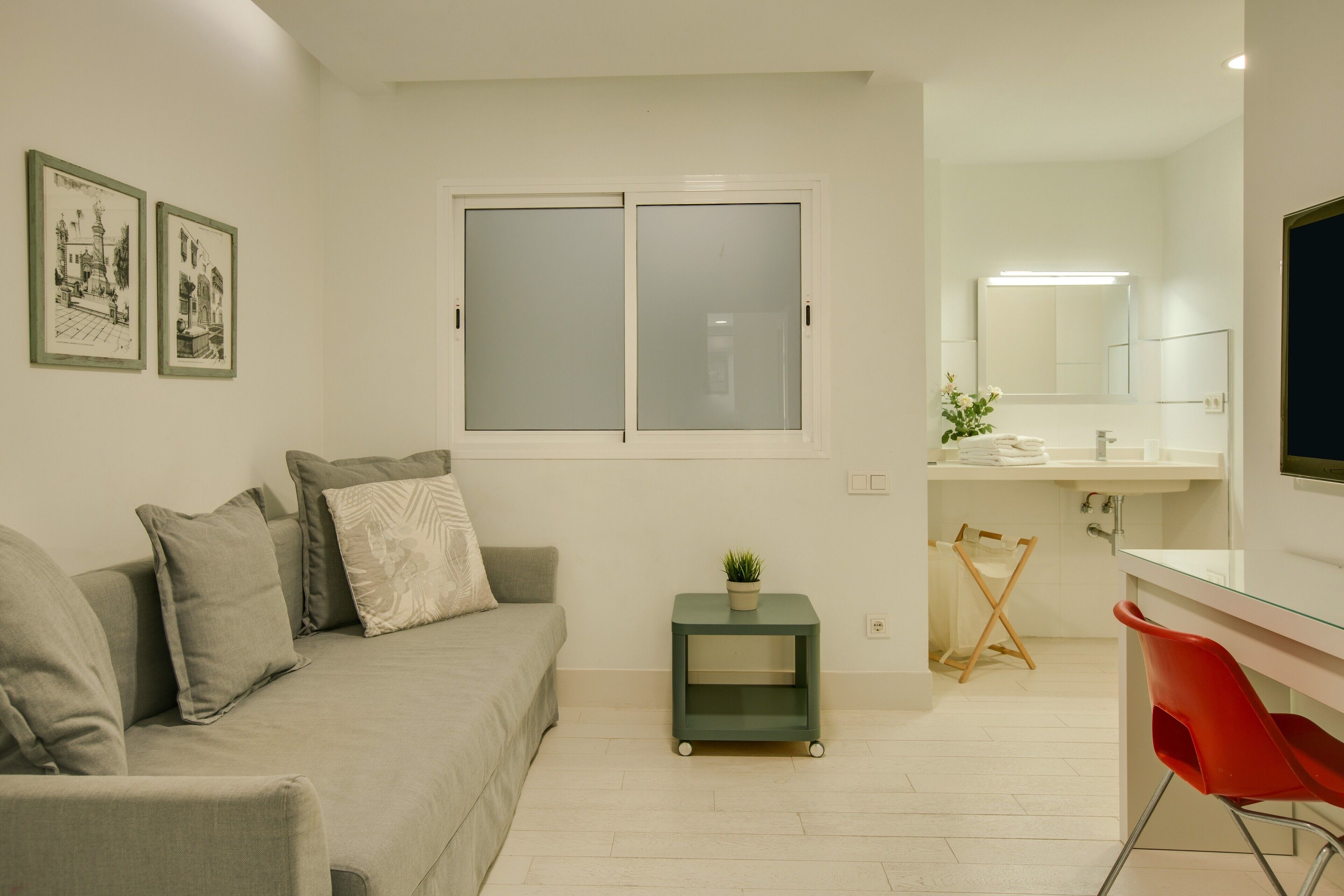 A modern living room with a sofa, a small side table with a plant, a window with frosted glass, and a bathroom with a mirror and sink.