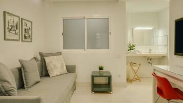 A modern living room with a sofa, a small side table with a plant, a window with frosted glass, and a bathroom with a mirror and sink.