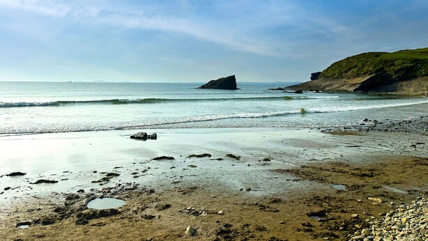 A beach with rocky terrain, calm water, and a distant rocky outcrop.