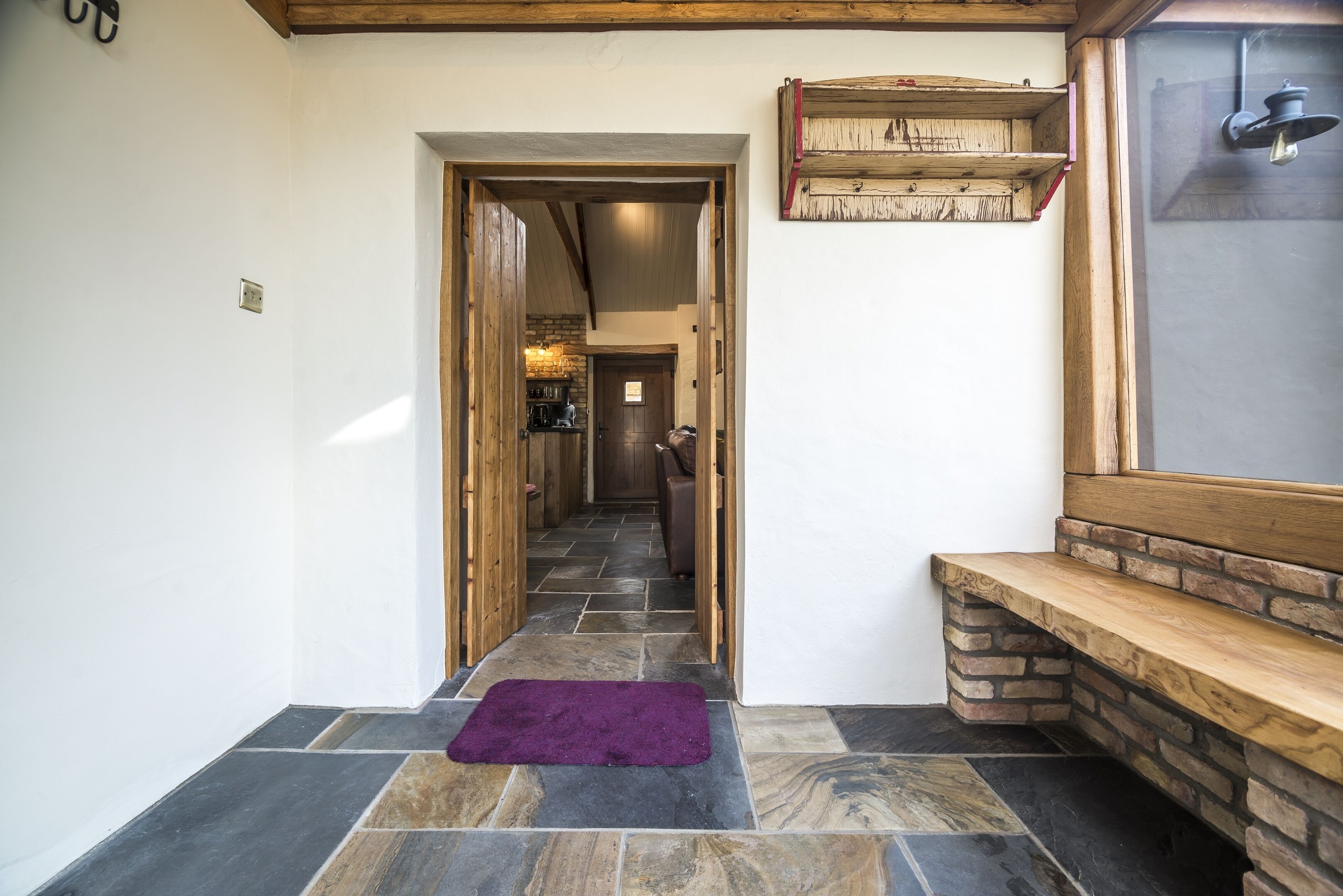 A hallway with stone flooring, a purple rug, wooden benches, and a rustic wall shelf.