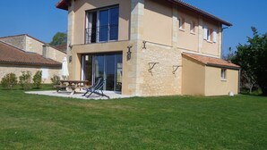 Una casa de dos pisos con fachada de piedra, ventanas grandes y un patio con mesa y sillas.