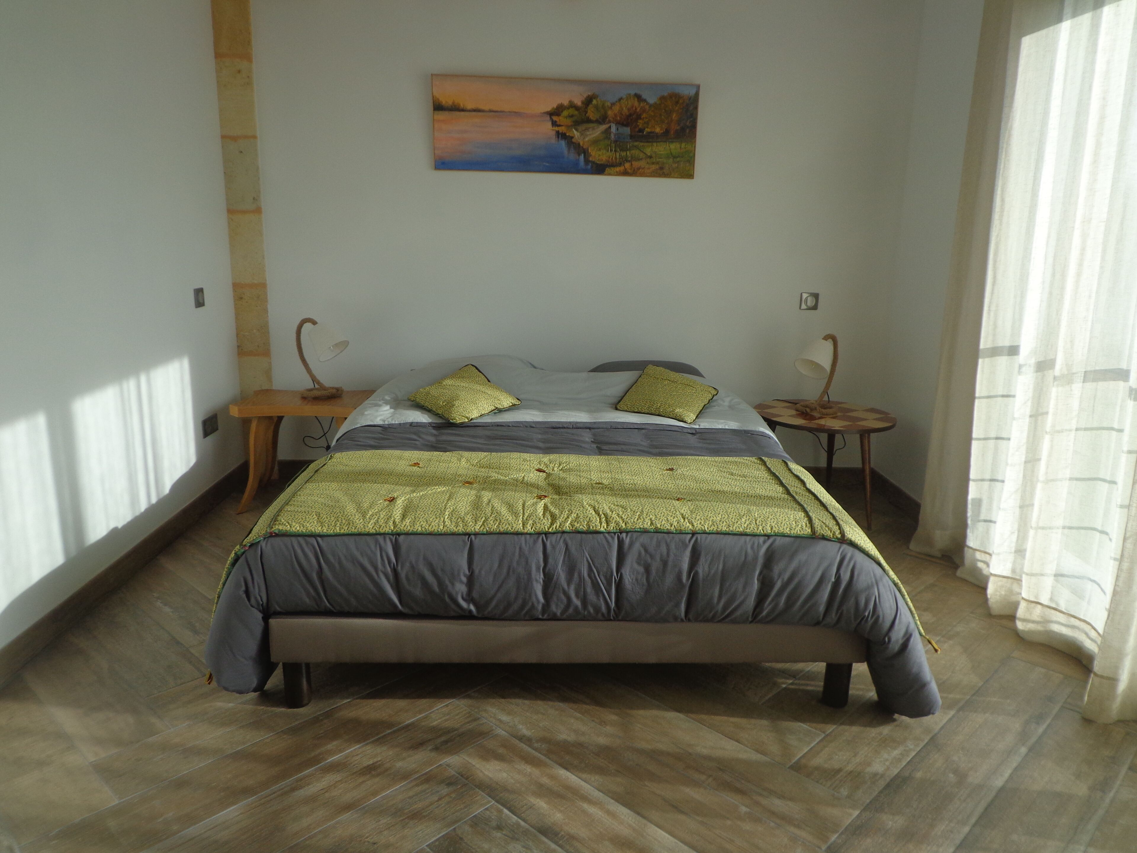 Un dormitorio con cama, dos mesitas de noche, lámpara, un cuadro en la pared y ventana con cortinas.