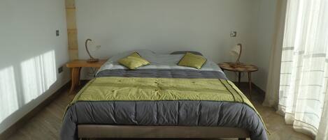 Un dormitorio con cama, dos mesitas de noche, lámpara, un cuadro en la pared y ventana con cortinas.