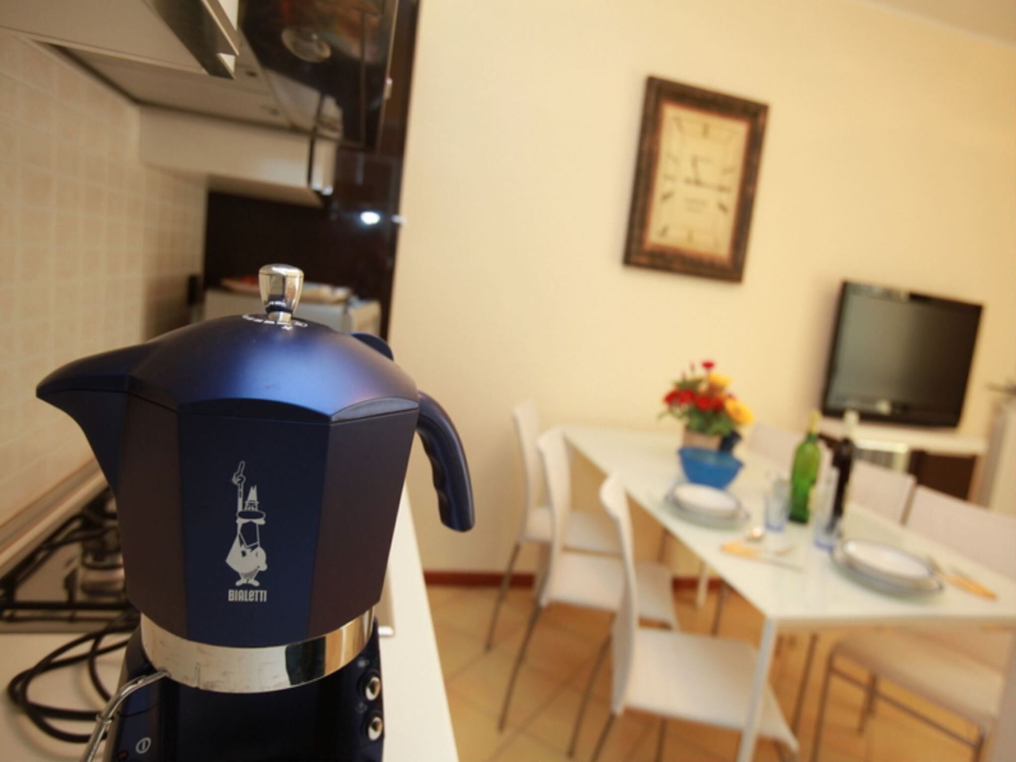 A kitchen with a coffee maker, a dining table with chairs, a TV, and a clock on the wall.