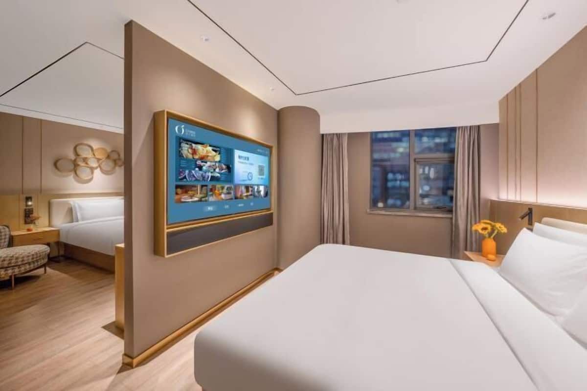 A hotel room with a large bed, a wall-mounted TV, a small table with a vase, and a window with curtains.