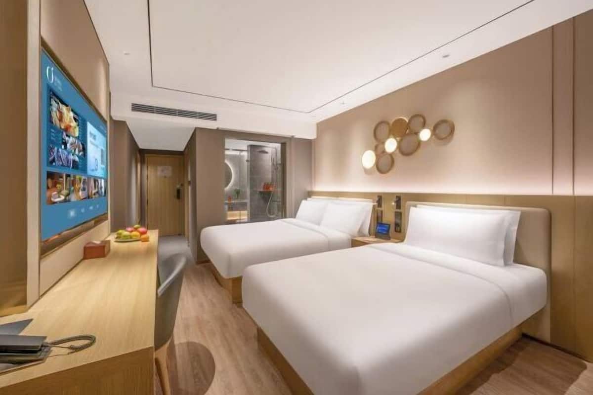 A modern hotel room with two beds, a desk with a laptop, and a wall-mounted TV displaying a menu.