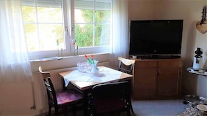 Un pequeño comedor con una mesa, sillas, un televisor y un ventanal con cortinas.