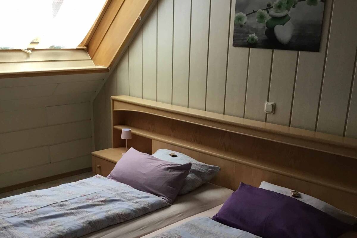 A dormitory room with two beds, a skylight, a painting on the wall, and a small bedside table.