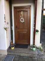 Uma porta de madeira com uma coroa de flores e uma placa de boas-vindas. Há duas plantas em vasos nos degraus e uma grade de metal em frente à porta.