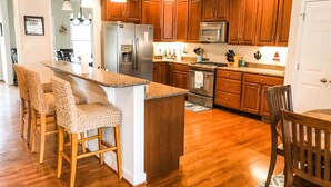 A modern kitchen with wooden cabinets, stainless steel appliances, and a central island with bar stools. - Coco’s Cabana (Deltaville)