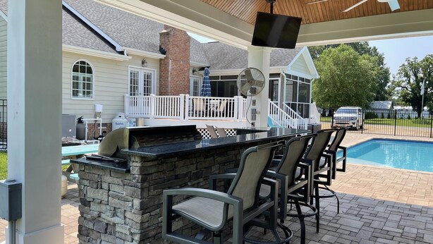 An outdoor bar area with stone accents, a pool, and a covered seating area with a ceiling fan. - Coco’s Cabana (Deltaville)