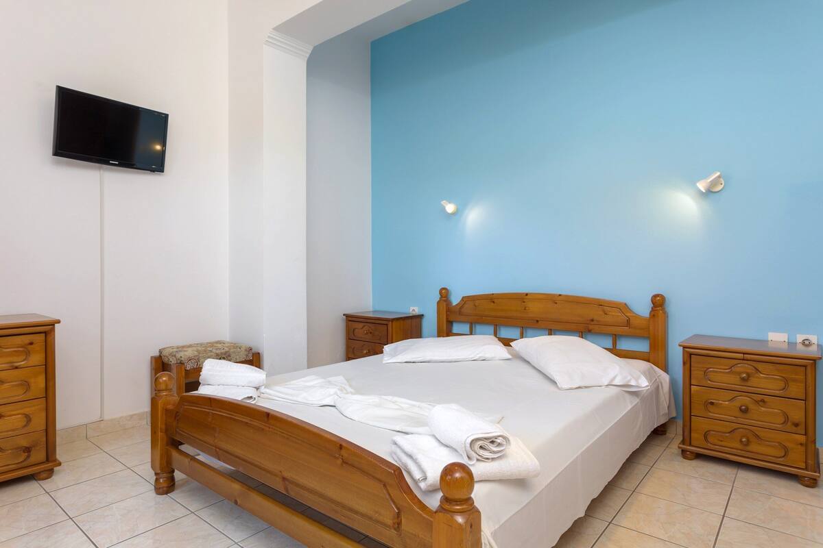 A bedroom with a wooden bed, white linens, a flat-screen TV on the wall, and a nightstand with drawers.