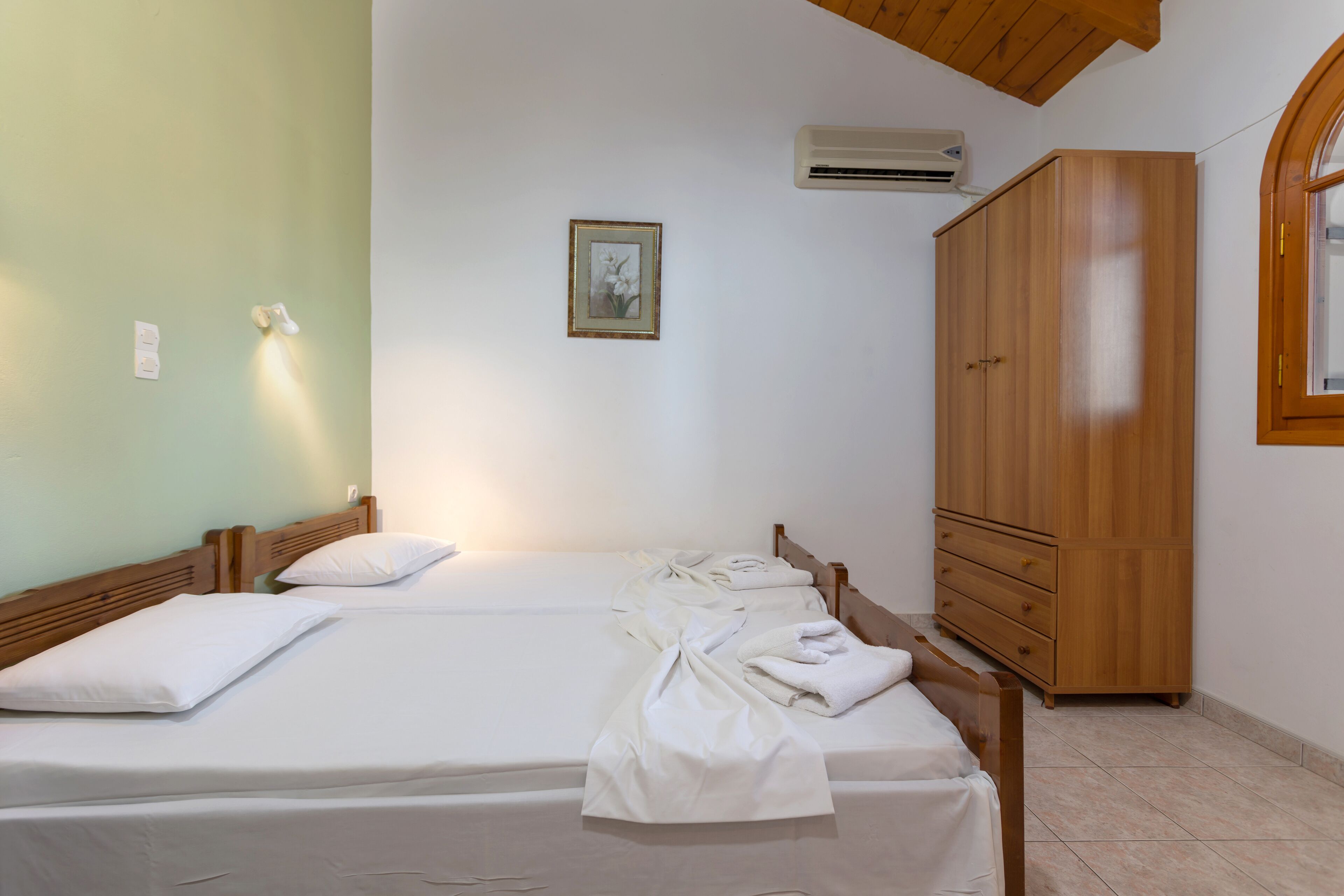 A bedroom with two beds, a wooden wardrobe, an air conditioning unit, and a framed picture on the wall.