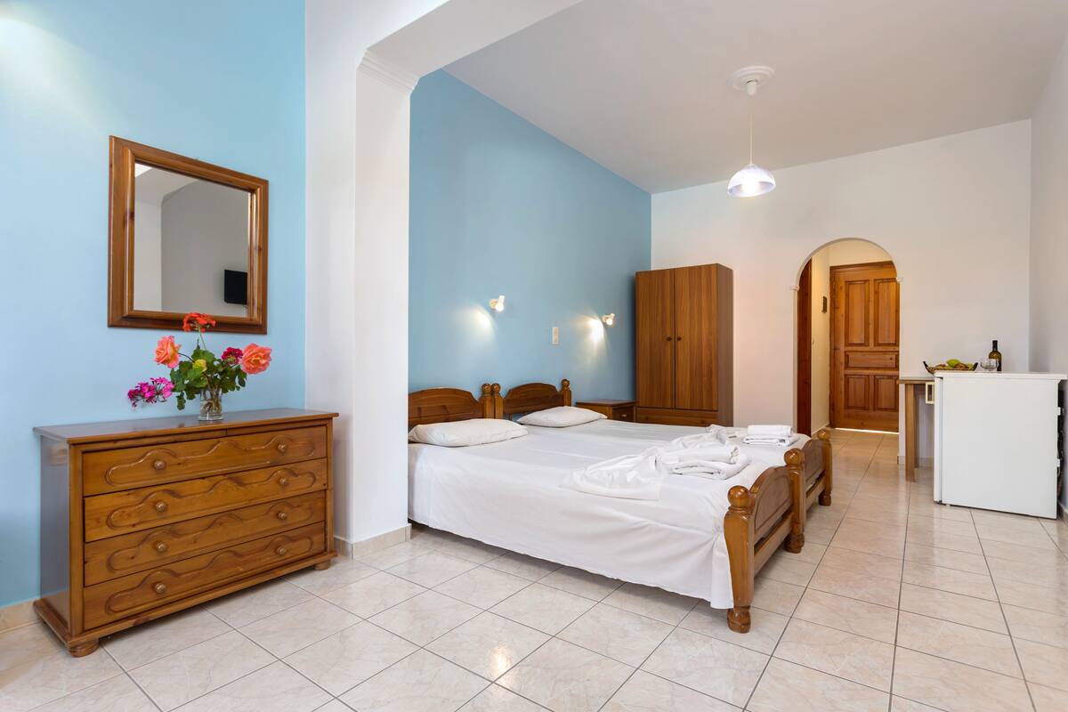 A bedroom with a wooden dresser, two beds, a mirror, and a small kitchenette.