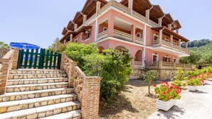 A multi-story building with a pink facade, a balcony on the second floor, and a staircase leading to the entrance. - Anemomylos Studios (Zakynthos)